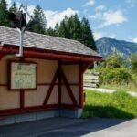 An idyllic alpine bus stop surrounded by lush greenery and mountains on a sunny day.