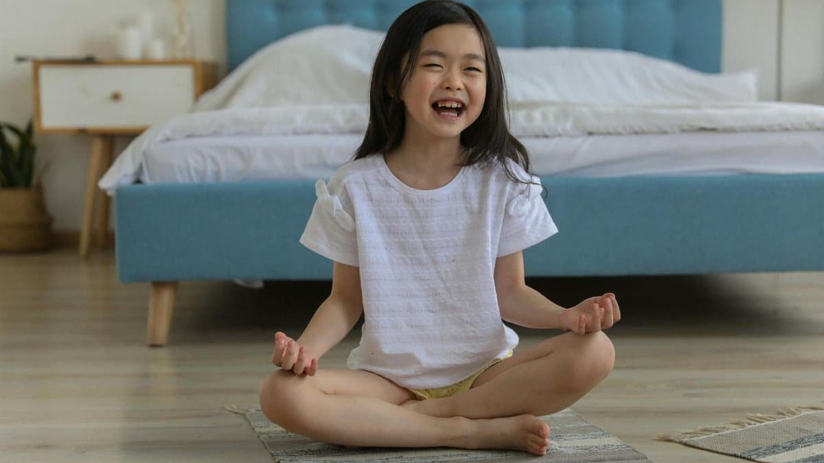 A cheerful young girl practicing yoga indoors, seated cross-legged and smiling brightly in a cozy bedroom.