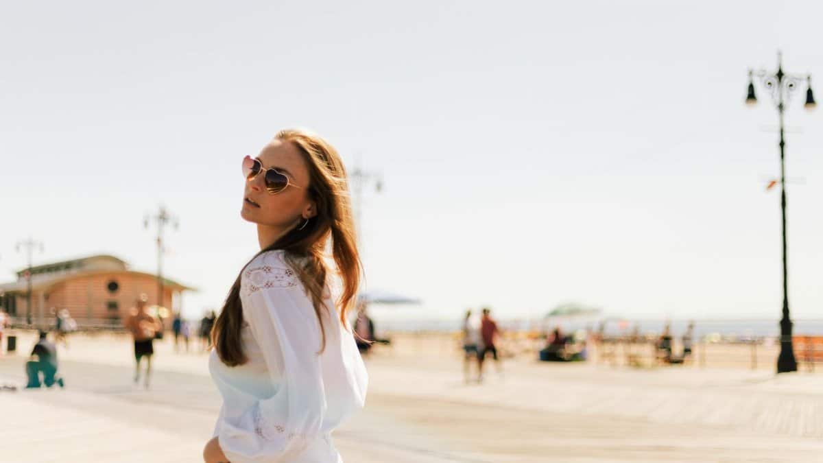 A fashionable woman wearing sunglasses leisurely strolls along Coney Island boardwalk under clear skies.