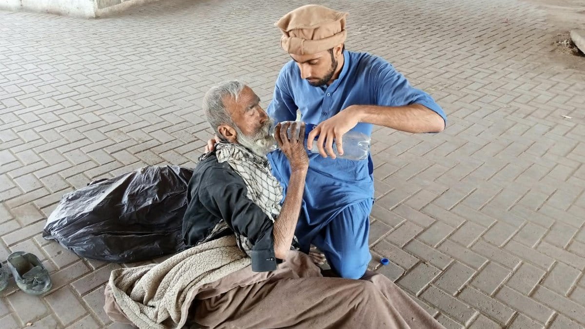 A young man helps a homeless senior by giving water outdoors in Karachi, Pakistan.