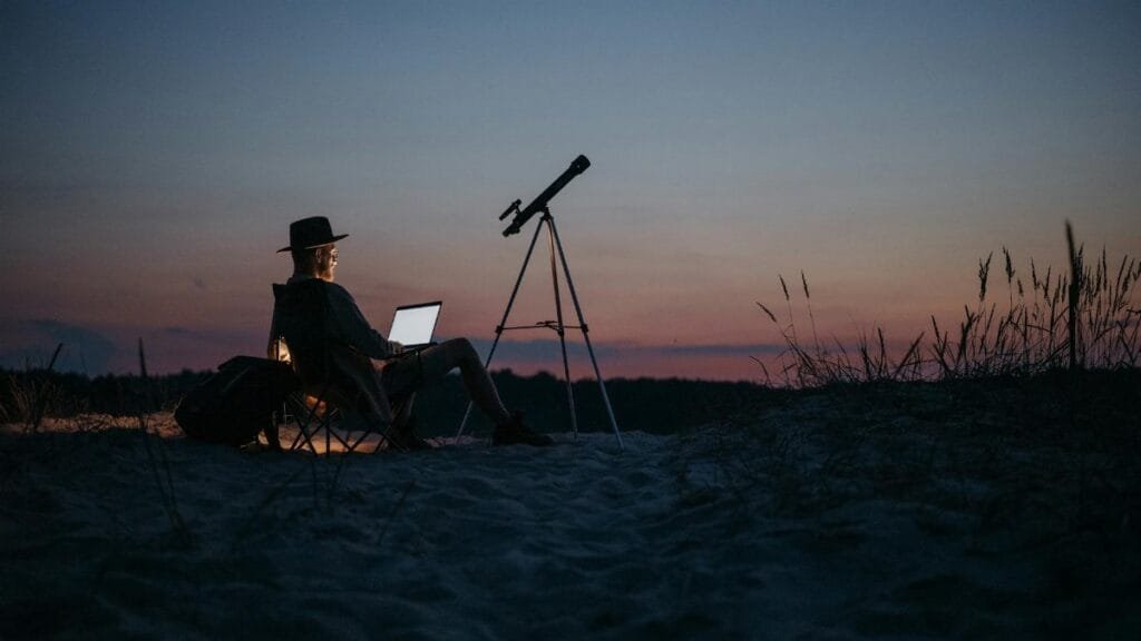 Person using a laptop and telescope at twilight, combining work and stargazing in a serene outdoor setting.