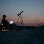 Person using a laptop and telescope at twilight, combining work and stargazing in a serene outdoor setting.