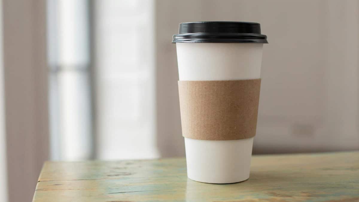 White paper coffee cup with brown cardboard sleeve on a rustic table indoors.