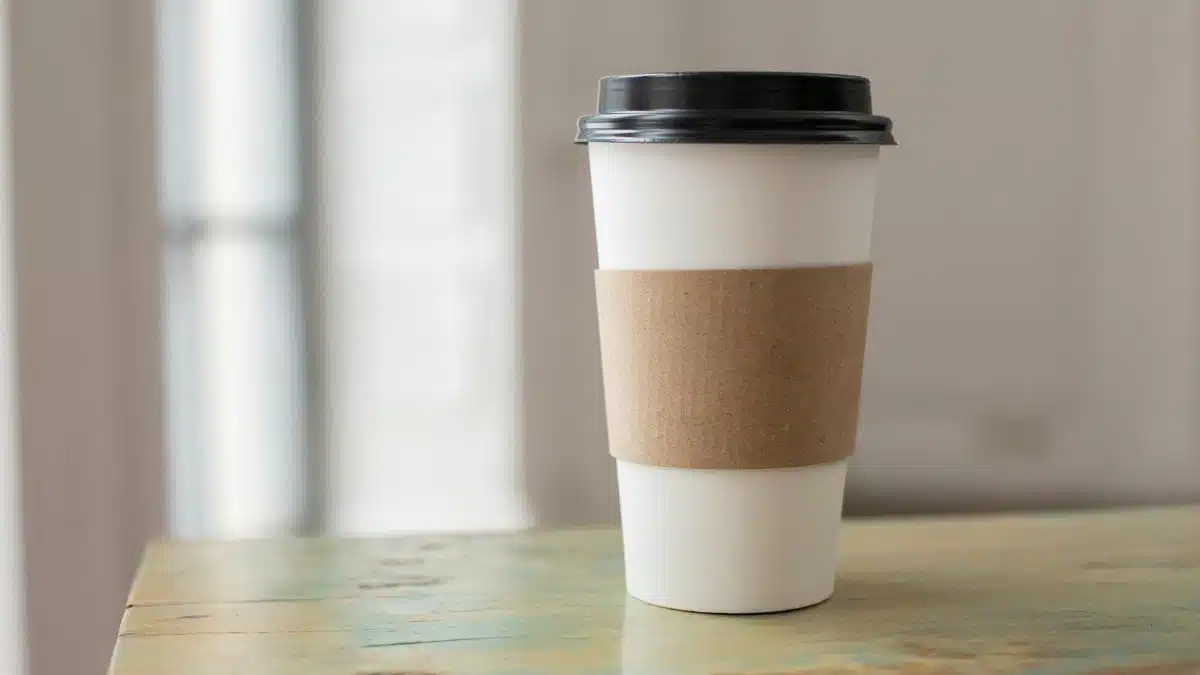 White paper coffee cup with brown cardboard sleeve on a rustic table indoors.