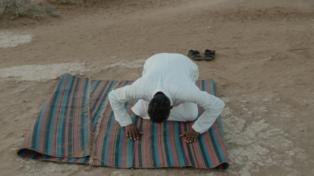 A man in traditional attire performs a prayer outdoors on a colorful mat, showcasing cultural beliefs.