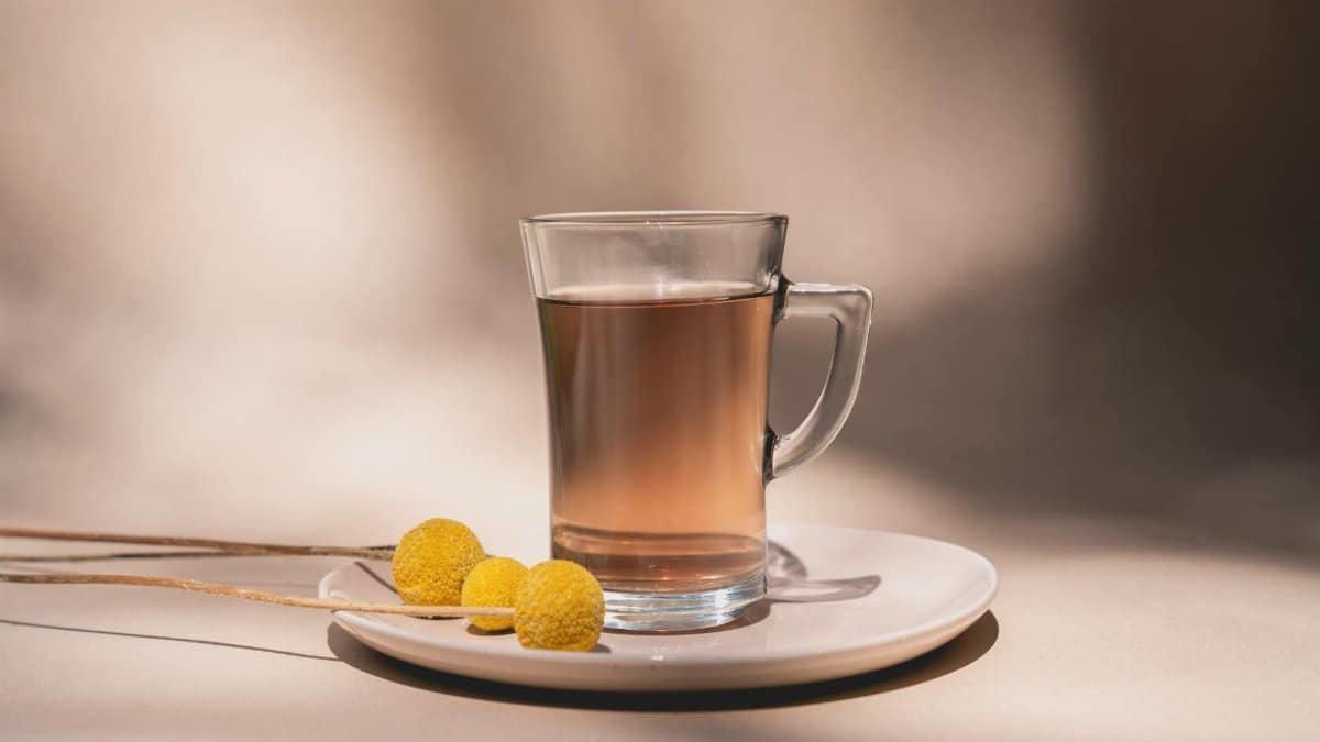 A clear tea glass with brown tea and yellow flowers on a white saucer in soft lighting.