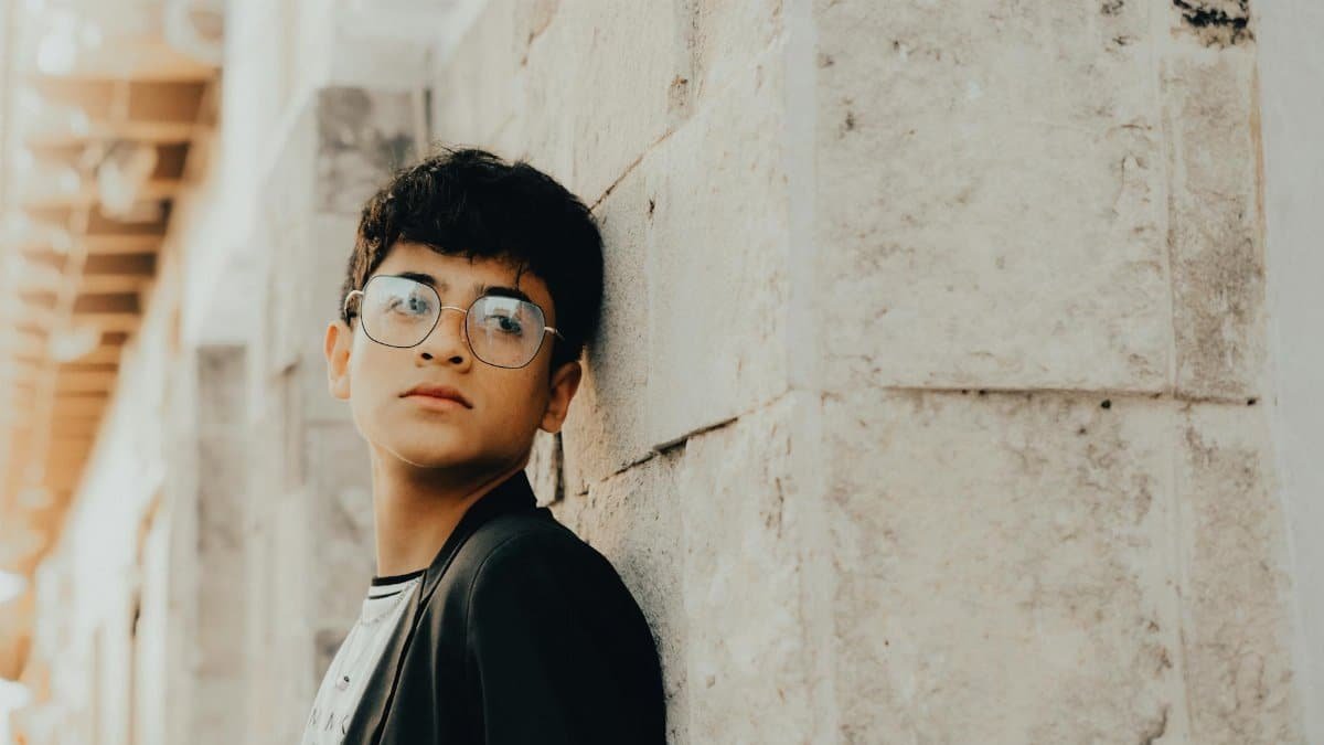 Portrait of a young boy with eyeglasses leaning against a stone wall outdoors.