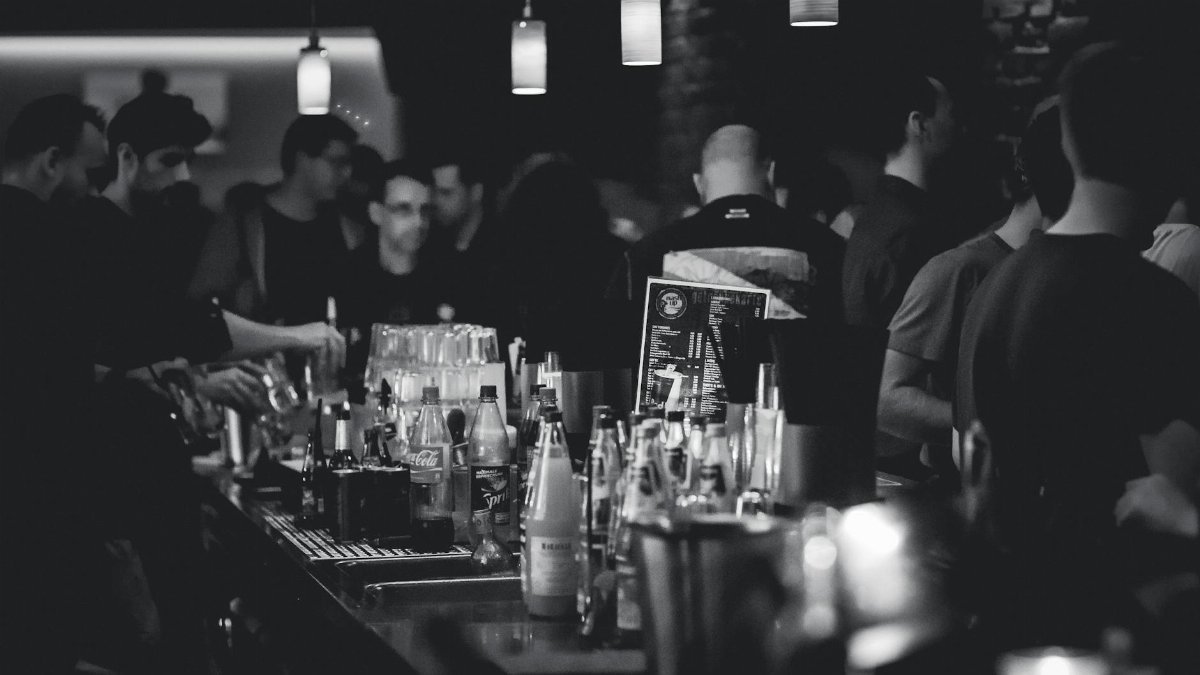 Black and white photo capturing the lively atmosphere in a bar with people socializing and a busy bartender serving drinks.
