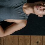 A man lies on his back in a yoga studio, relaxing on a mat in natural light.