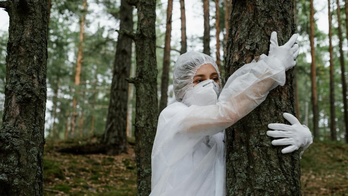 Individual in hazmat suit and face mask hugging tree, symbolizing connection with nature.