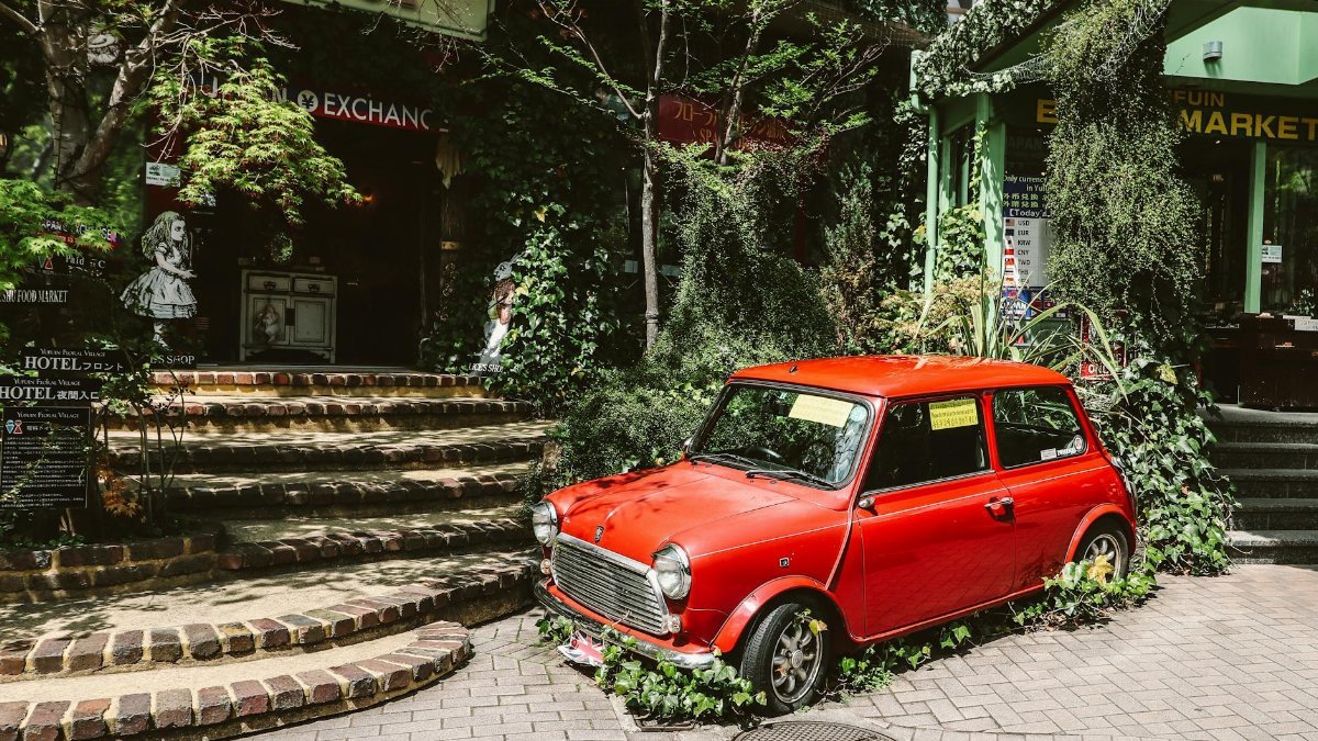 A classic red Mini Cooper parked amidst lush greenery and old town storefronts.