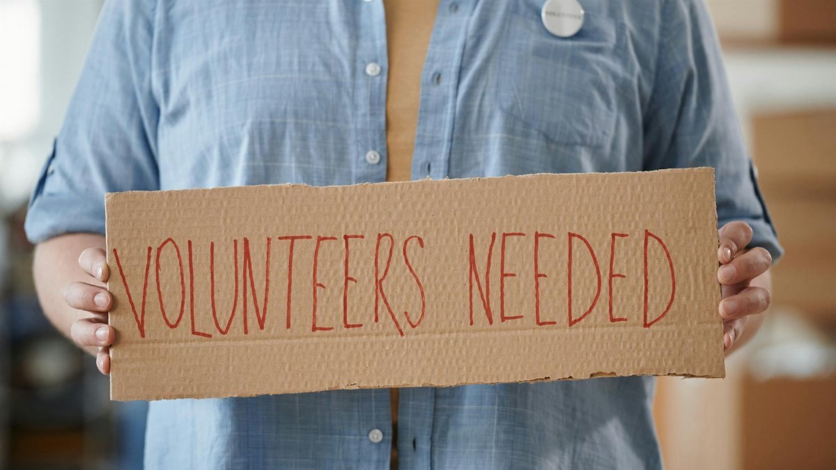 Person holding a sign reading 'Volunteers Needed' to encourage community support.