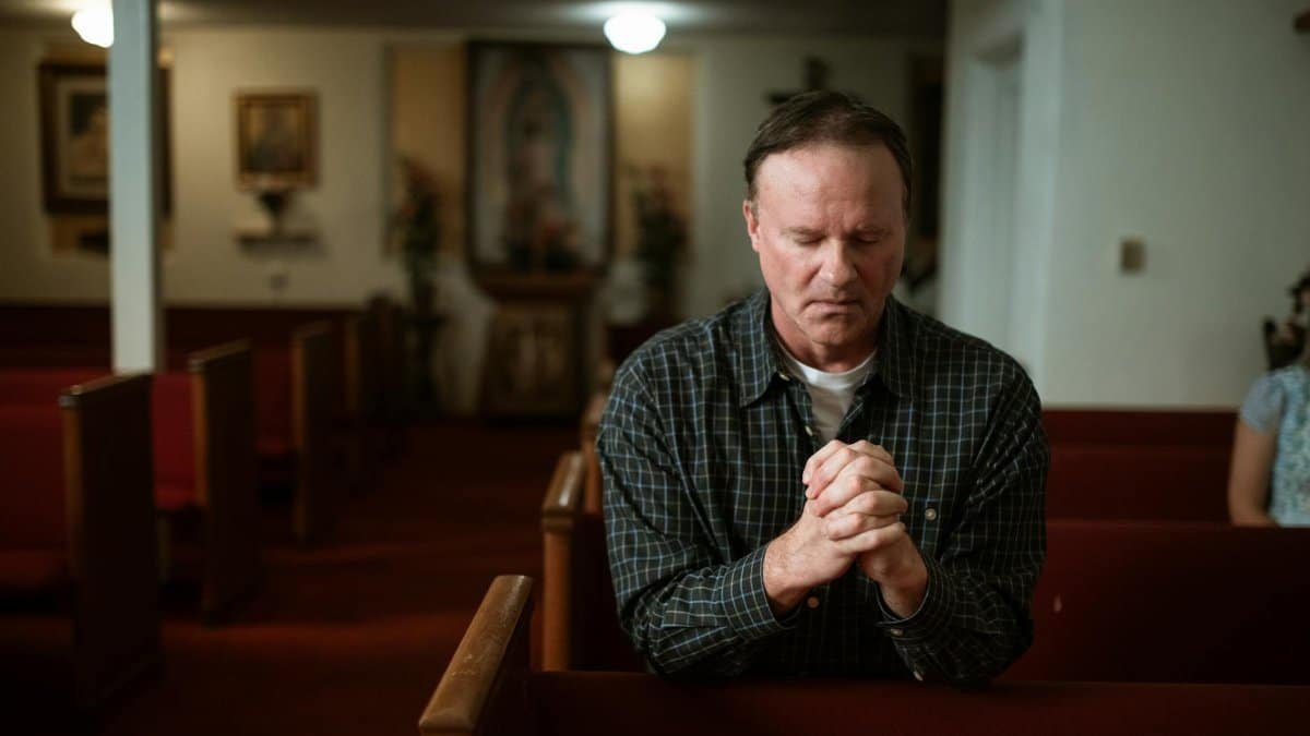Adult man kneeling in church pew, praying with devotion in a quiet, sacred setting.