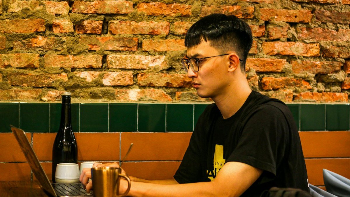 Young man studying on laptop in a cozy cafe setting with exposed brick wall and ambient lighting.
