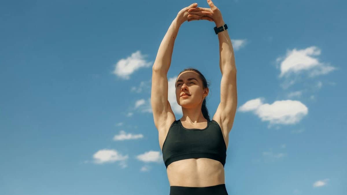 Woman in sports bra stretching under a clear blue sky, embodying fitness and wellness.