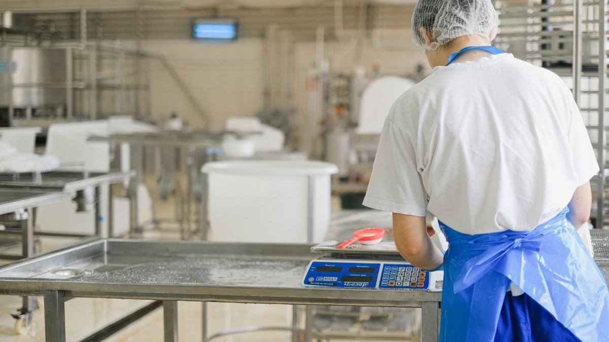 A factory worker in uniform measures ingredients on a digital scale in an industrial setting.