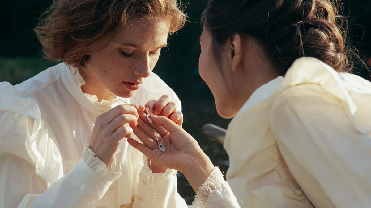 Two women share an intimate moment outdoors, holding a necklace together.