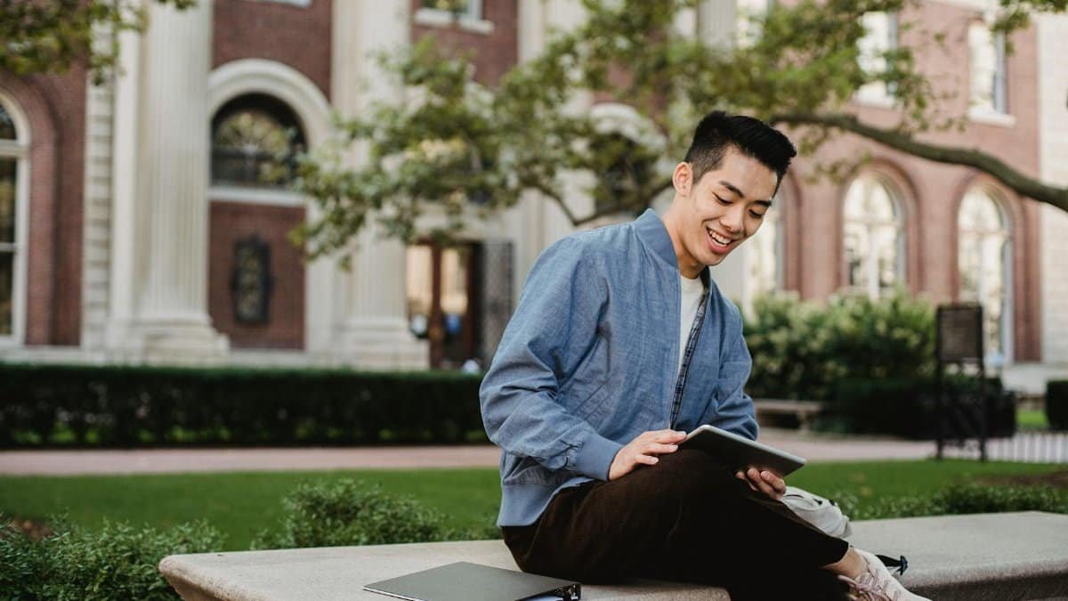 Young man joyfully using a tablet while seated outside a historic campus building.