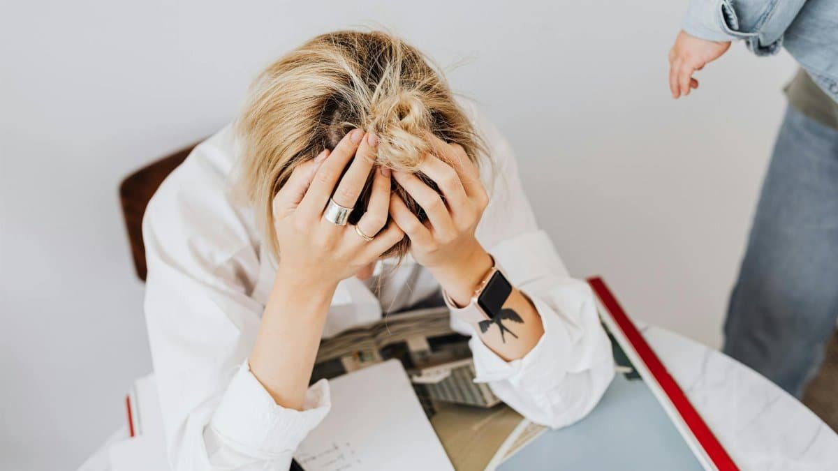 Frustrated woman in white shirt sitting with head in hands, overwhelmed by work tasks.
