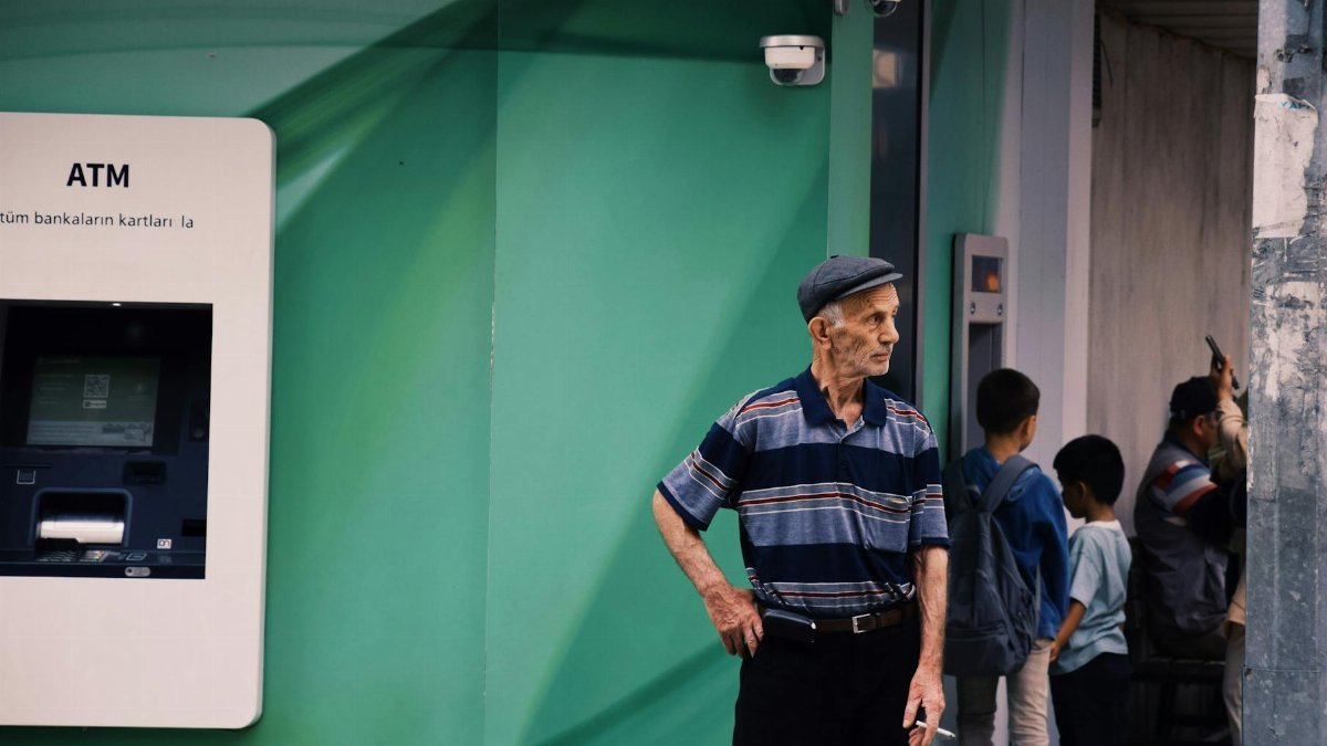 Elderly man in a striped shirt stands by an ATM with children nearby in an urban setting.