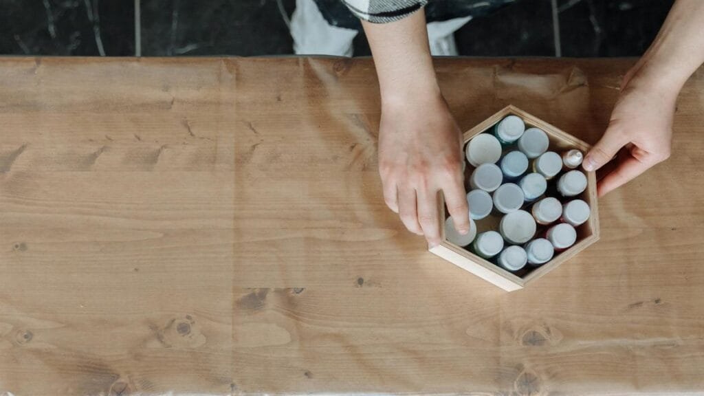 An artist's hands arranging paint bottles in a hexagonal box on a work table.