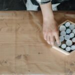 An artist's hands arranging paint bottles in a hexagonal box on a work table.
