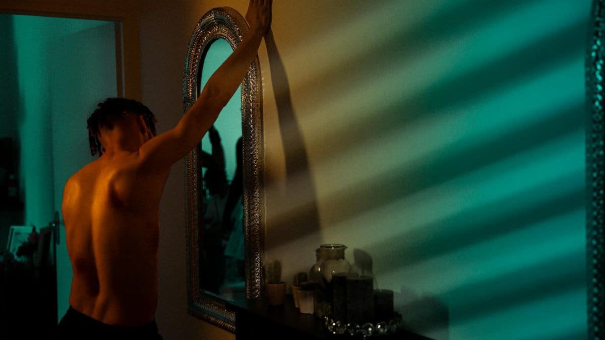A shirtless man reflects deeply in front of a mirror with dramatic lighting indoors.