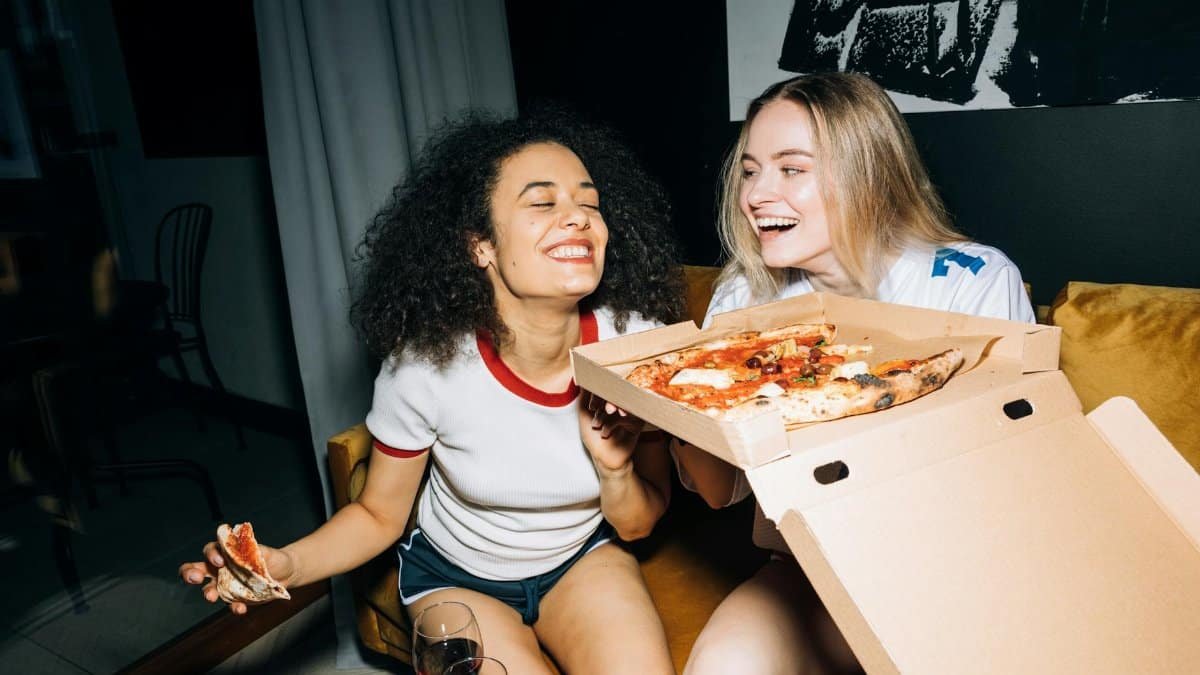 Two happy friends laughing and enjoying pizza indoors during a cozy night in.