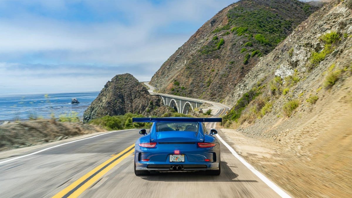 Blue sports car drives along a scenic coastal highway, hugging cliffs with the sea in view.