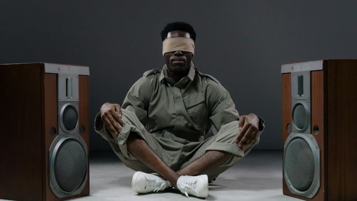 Blindfolded man in green outfit sitting between large speakers in a minimalist setting.