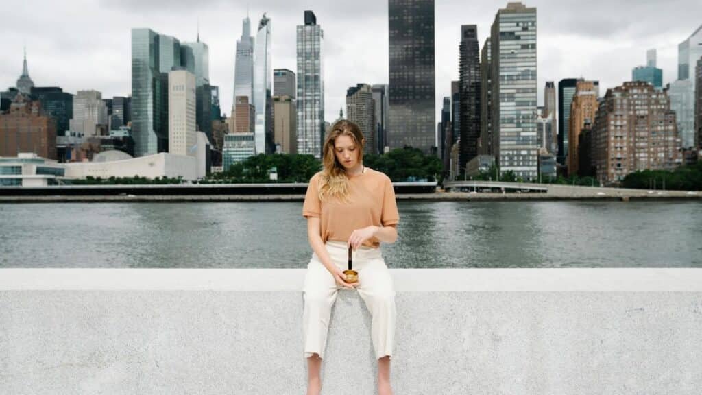 A woman meditates with a singing bowl in front of the New York City skyline.
