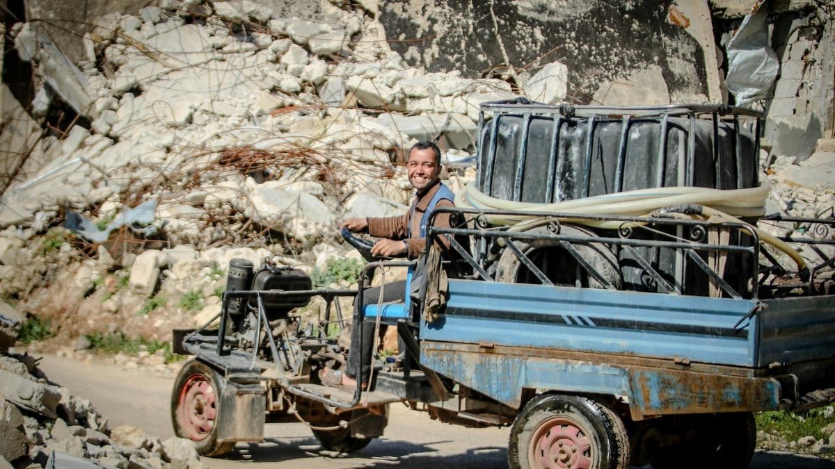 Smiling man driving a makeshift vehicle through rubble-strewn street in Idlib, Syria under bright sunlight.