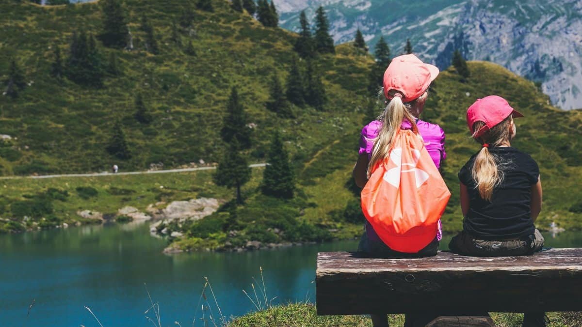 Two young girls sitting on a bench overlooking a serene mountain lake, enjoying a summer hike.