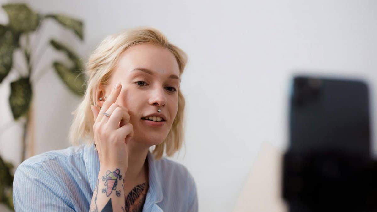 A young woman with tattoos recording a makeup tutorial with a smartphone indoors.