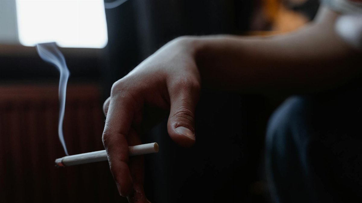 Close-up of a hand holding a lit cigarette indoors with smoke rising in a dimly lit room.