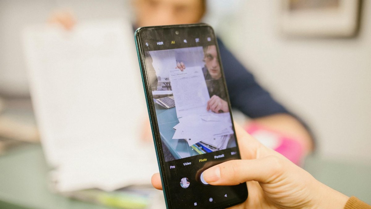 Close-up of student using smartphone to photograph examination papers in a classroom setting.
