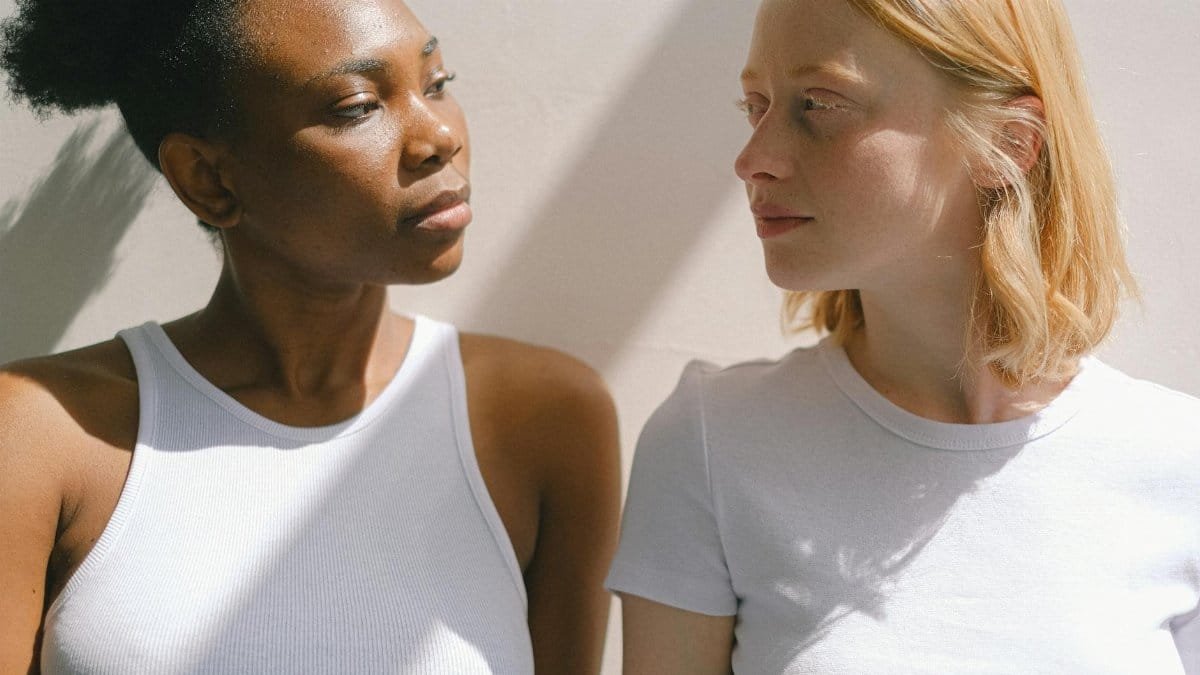 Two diverse women in white tops share a moment of connection in soft daylight.