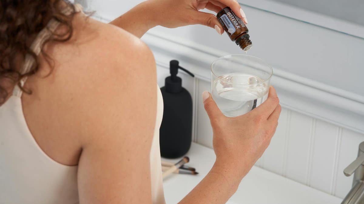 A woman adds essential oil to a glass of water in a bathroom setting, emphasizing wellness.