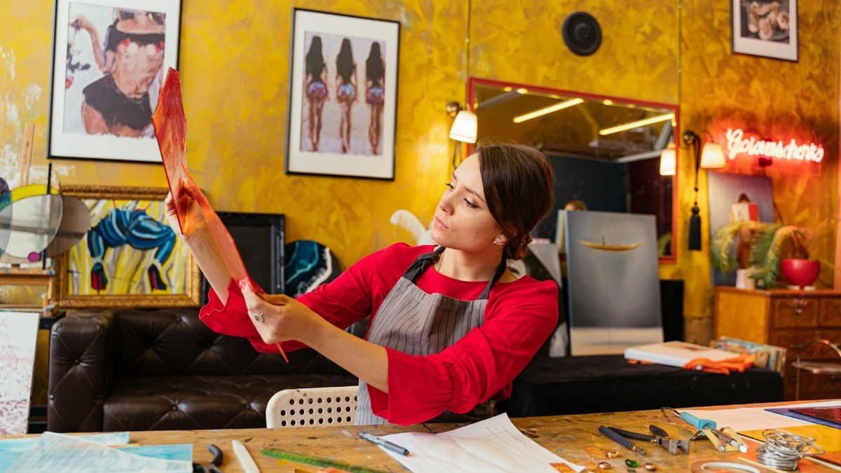 Artist wearing an apron examines her artwork in a colorful, art-filled studio.