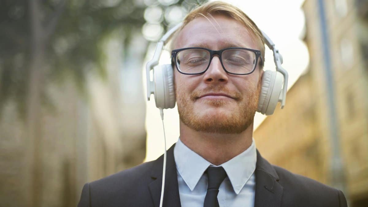 Portrait of a young man in a suit, wearing headphones and enjoying music outdoors with a blurred urban background.