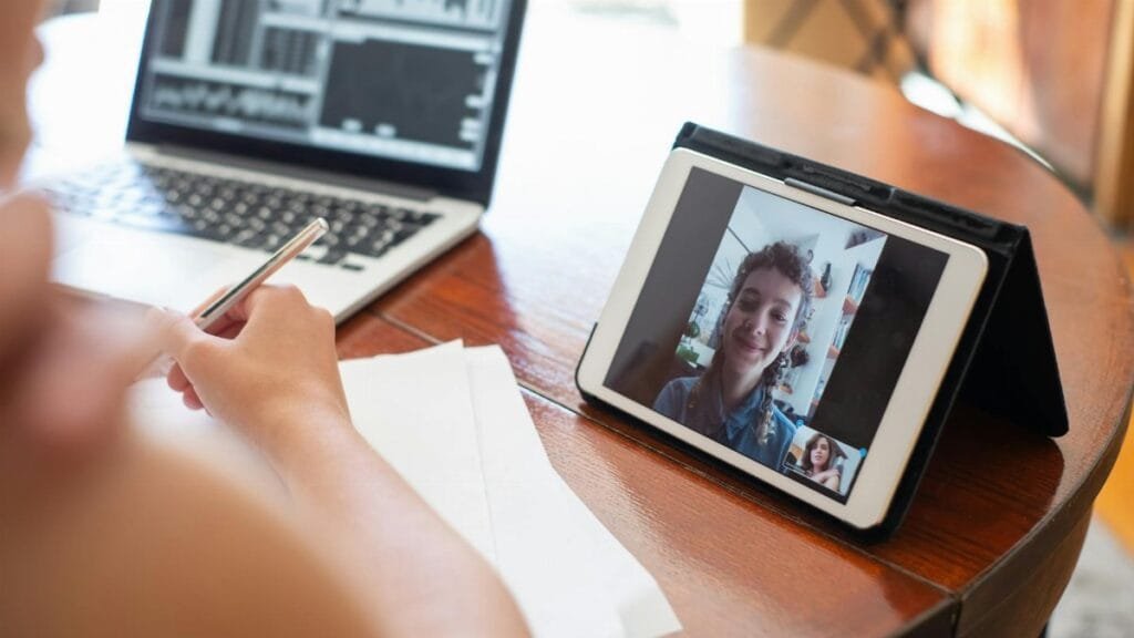 A person on a video call using a tablet, with a laptop displaying data nearby.
