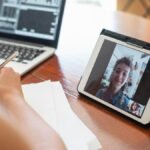 A person on a video call using a tablet, with a laptop displaying data nearby.