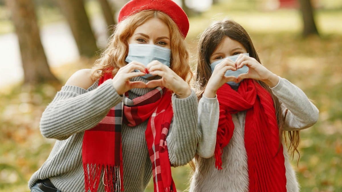 Mother and daughter making heart symbols in a park wearing masks, expressing love and safety.