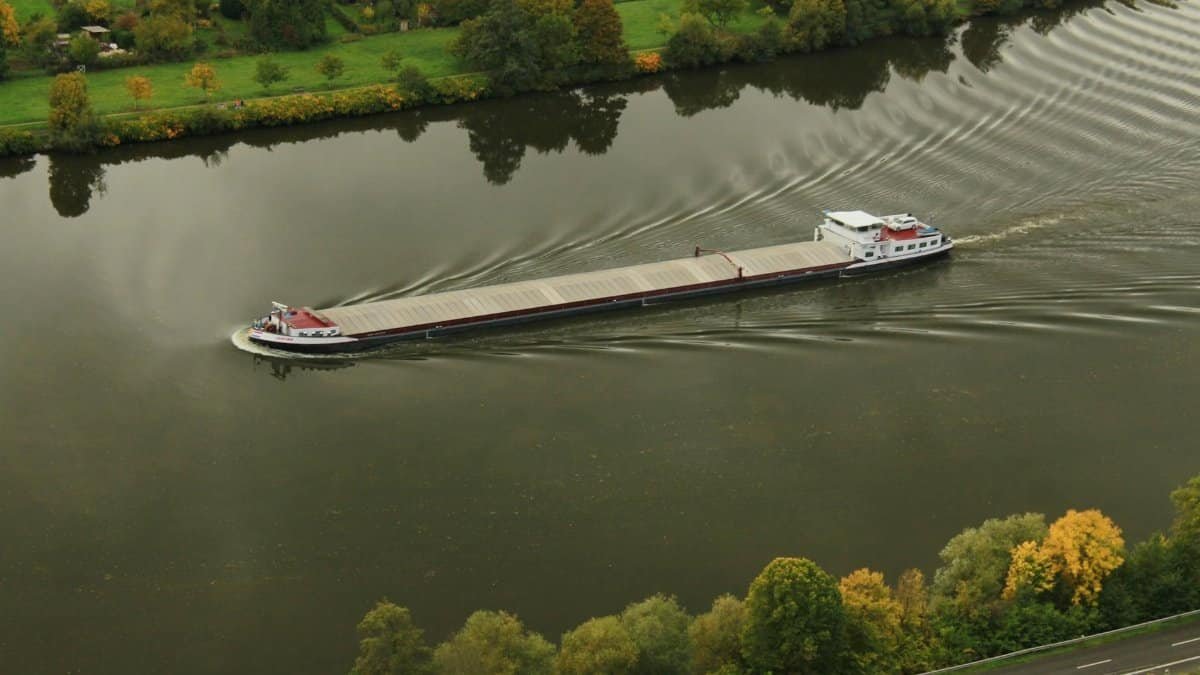 A large industrial ship navigating a scenic river bend, surrounded by lush autumn foliage.