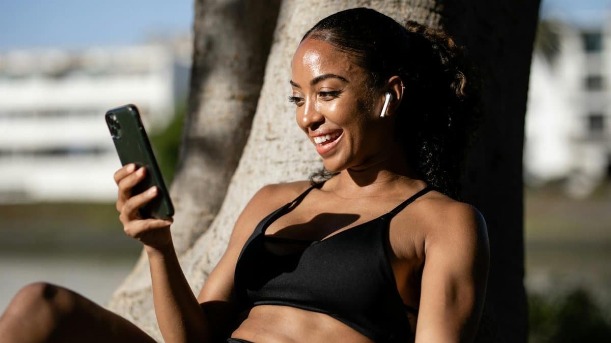 Smiling woman in sportswear using smartphone with earbuds, enjoying the sunny outdoors.