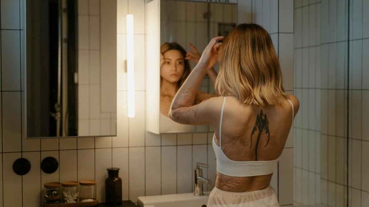 A woman with tattoos and burn scars looks at her reflection in a bathroom mirror.