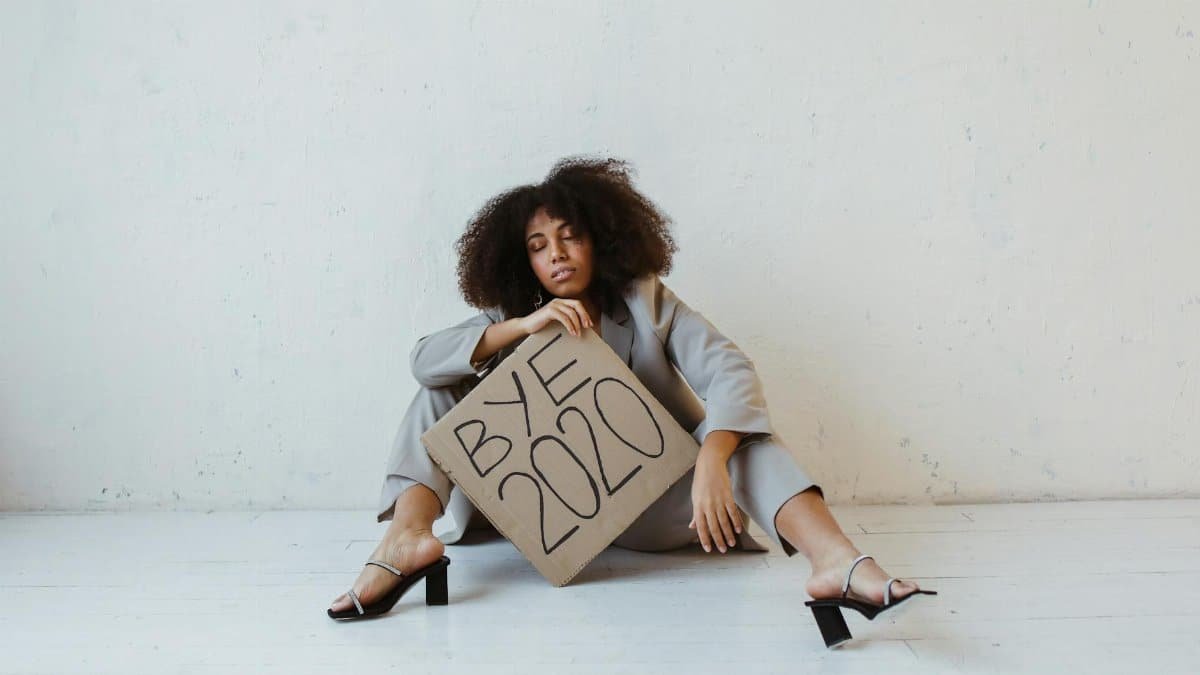 Woman sitting on floor holding 'Bye 2020' sign, expressing exhaustion and relief.