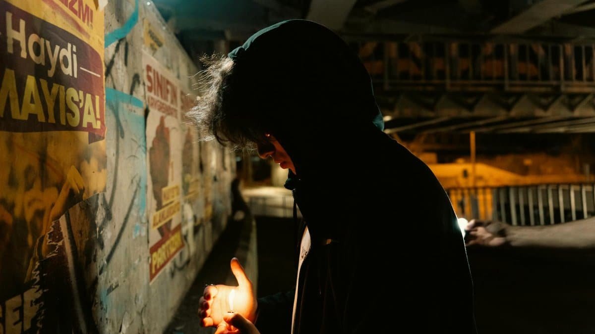 Hooded figure holding a light source at night in an urban underpass.