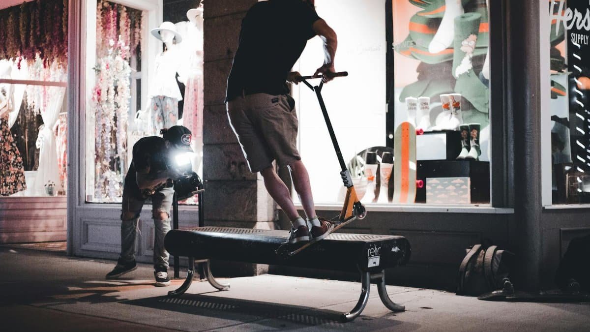 A videographer captures a scooter trick on a bench in front of an illuminated storefront at night.