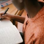 Focused black woman writing in a notebook with stacked books on a desk, illustrating learning.